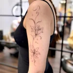 Zartes Fine Line Tattoo mit Lilienblüten, geschwungenen Ranken und Funkel-Details auf dem Oberarm.