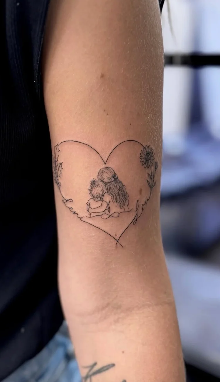 Zartes Fineline Herz Tattoo am Oberarm mit einer liebevollen Mutter-Kind-Darstellung, floralen Details und feiner Linienführung – persönliches Tattoo in Berlin.