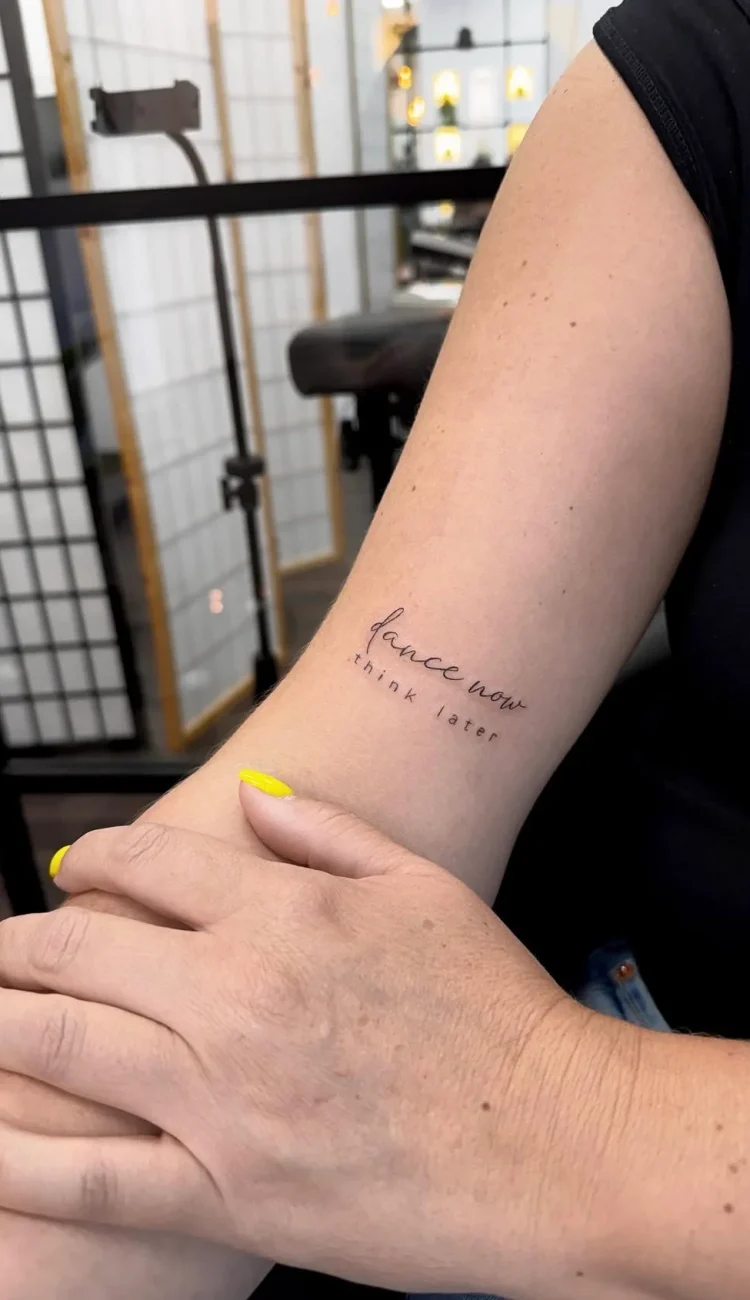 Filigranes Fineline Schriftzug Tattoo mit dem Spruch „dance now think later“ am Arm – minimalistisches Lettering Tattoo mit feiner Linienführung in Berlin.