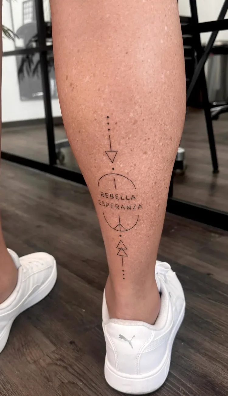 Vertikales geometrisches Tattoo mit dem Schriftzug Rebella Esperanza und minimalistischen Symbolen an der Wade.