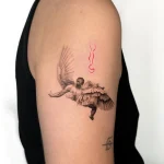 Filigranes Microrealismus Tattoo von Ikarus mit gefiederten Flügeln und rotem Farbakzent am Oberarm, gestochen in einem Berlin Tattoo Studio.