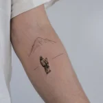 Detailliertes Miniatur Tattoo eines Snowboarders vor einer Bergkulisse in feinen Linien am Arm.